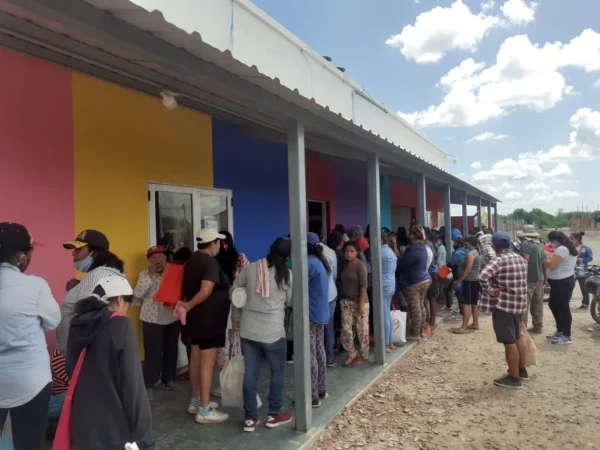 Una fila larga de personas esperando frente a un edificio de un solo piso con paredes pintadas de bloques de colores vibrantes (rosa, amarillo y azul). El lugar tiene un alero sostenido por columnas metálicas y se encuentra en un entorno de suelo de tierra bajo un cielo parcialmente nublado.