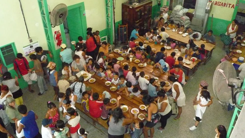 Vista aérea de un comedor comunitario lleno de niños y adultos sentados en mesas largas de madera durante la hora del almuerzo.