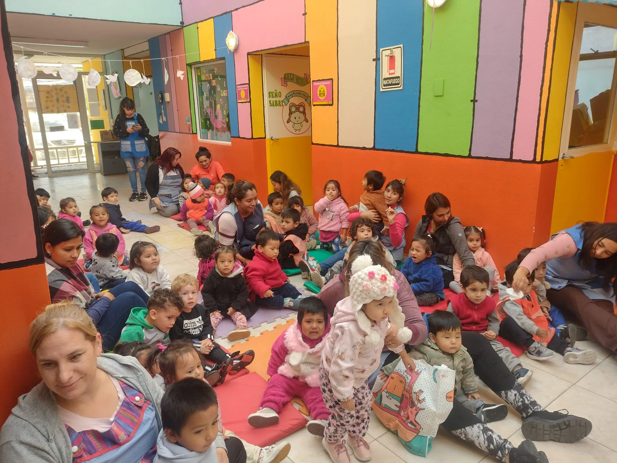 Un grupo numeroso de niños pequeños y maestras sentados en el suelo de un jardín de infantes colorido. Las paredes del fondo tienen rayas verticales de colores vibrantes y una puerta con el cartel 'Sala Verde'. Algunos niños visten ropa de abrigo y gorros, mientras participan en una actividad grupal sobre colchonetas de colores.