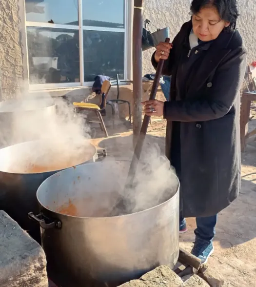 Una mujer revolviendo una olla popular humeante con una cuchara de madera larga en un espacio al aire libre