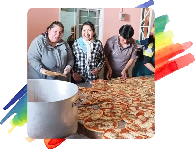 Tres personas preparando una gran cantidad de pizzas caseras en una mesa larga dentro de un comedor comunitario, con un ambiente alegre y una olla grande al frente