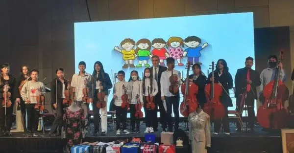 Un grupo de niños y jóvenes de una orquesta infantil posando en un escenario con sus instrumentos, incluyendo violines, chelos y un contrabajo. Detrás de ellos, una pantalla gigante muestra una ilustración infantil de cinco niños tomados de la mano. En la parte frontal del escenario se observan diversos objetos y prendas de vestir en exhibición.