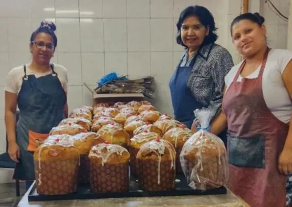 Tres mujeres con delantales posando con orgullo junto a una mesa llena de pan dulces recién horneados y decorados en un taller de panadería social.