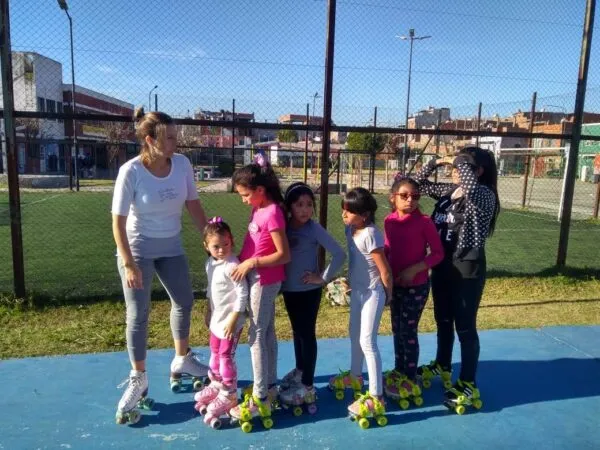 Una instructora y un grupo de niñas practicando patinaje artístico sobre una pista azul al aire libre. Todas llevan patines de cuatro ruedas y ropa deportiva. Al fondo se ve una cancha de césped sintético cercada por un alambrado y edificios bajos bajo un cielo despejado.