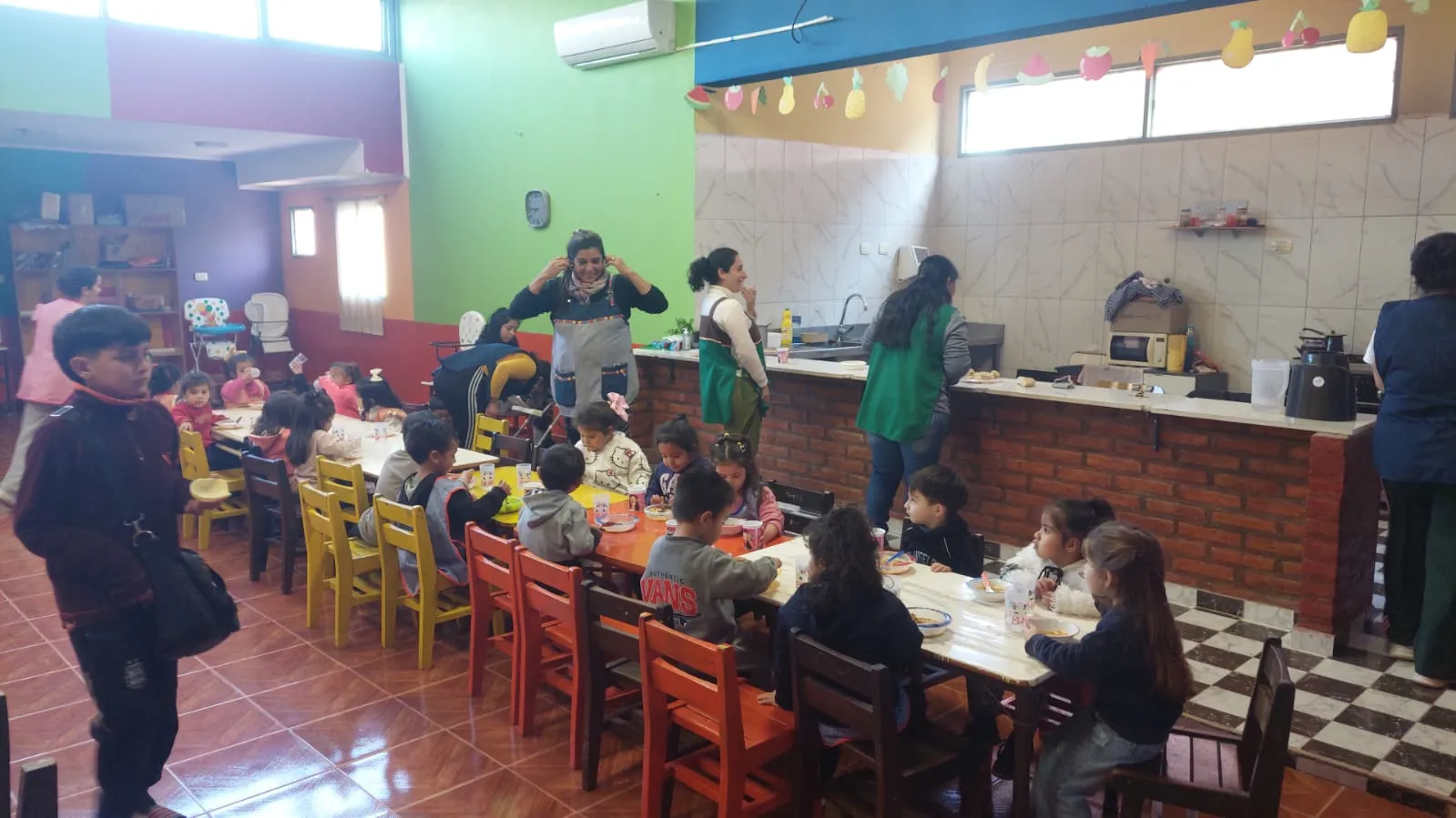 Sala de un centro de primera infancia con niños pequeños sentados en mesas largas de madera almorzando o merendando en sillas de colores, mientras maestras y personal con delantales los asisten cerca de una barra de ladrillos que conecta con la cocina.