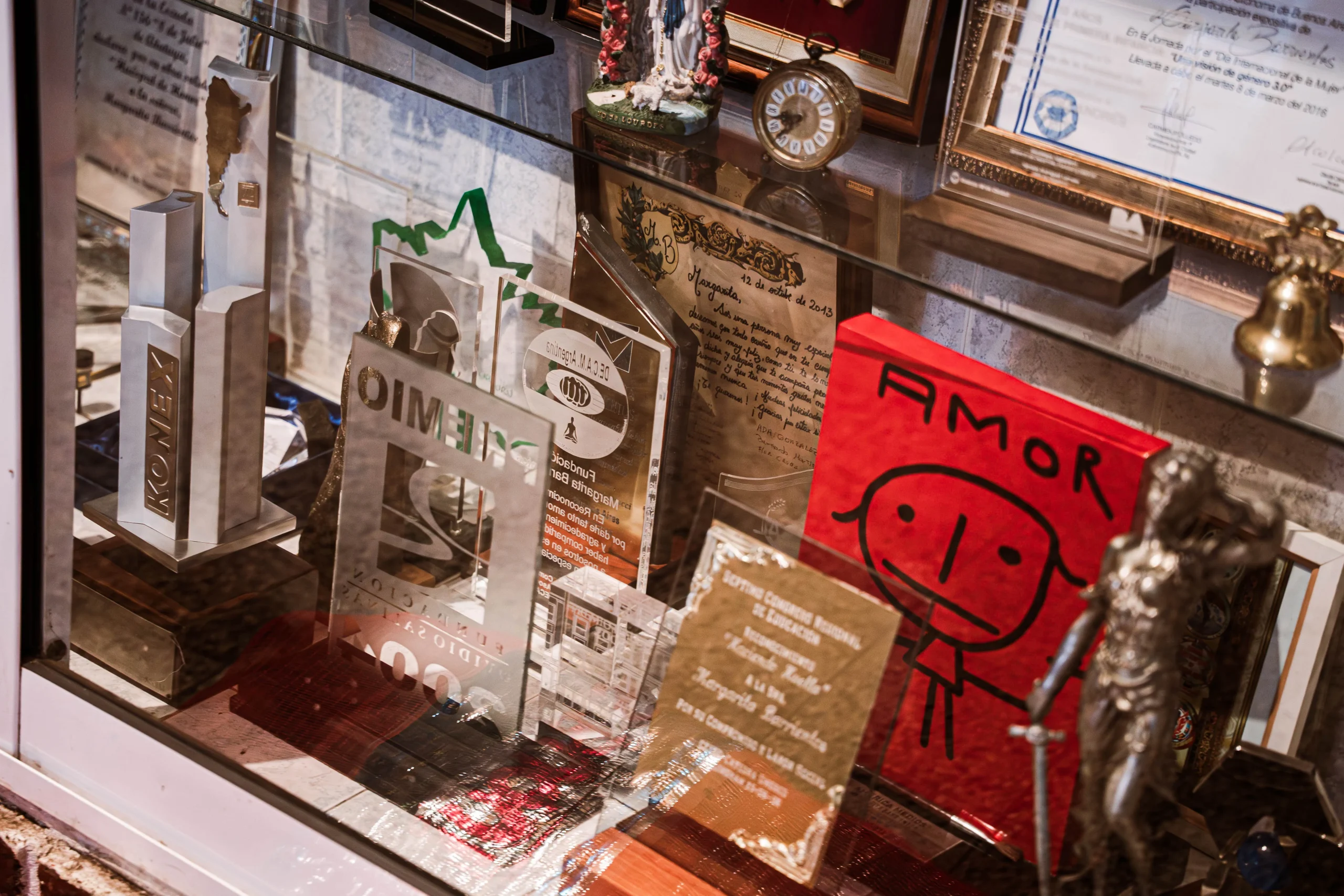 Primer plano de una vitrina de vidrio repleta de trofeos, placas conmemorativas y diplomas. Se distinguen premios de acrílico y metal, incluyendo uno con la inscripción 'KONEX'. También hay objetos decorativos como un reloj antiguo, una figura religiosa, una campana pequeña y un cartel rojo que dice 'AMOR' con un dibujo infantil