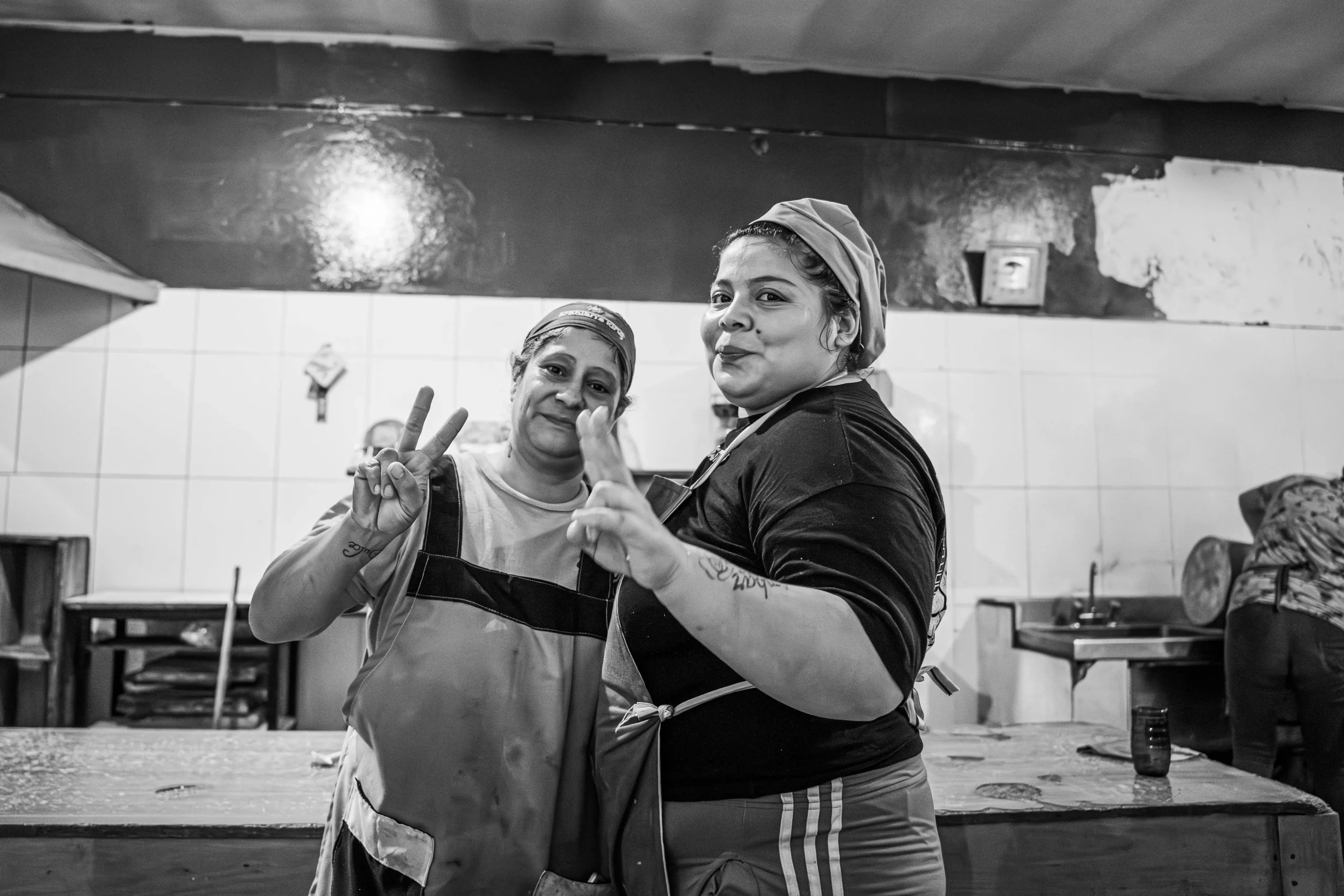 Fotografía en blanco y negro de dos mujeres cocineras posando sonrientes y de pie en una cocina amplia. Ambas están haciendo el gesto de 'v de victoria' o paz con sus manos. Llevan delantales de trabajo y gorros para el cabello. Al fondo se ve la estructura de la cocina con azulejos, estantes y ollas.