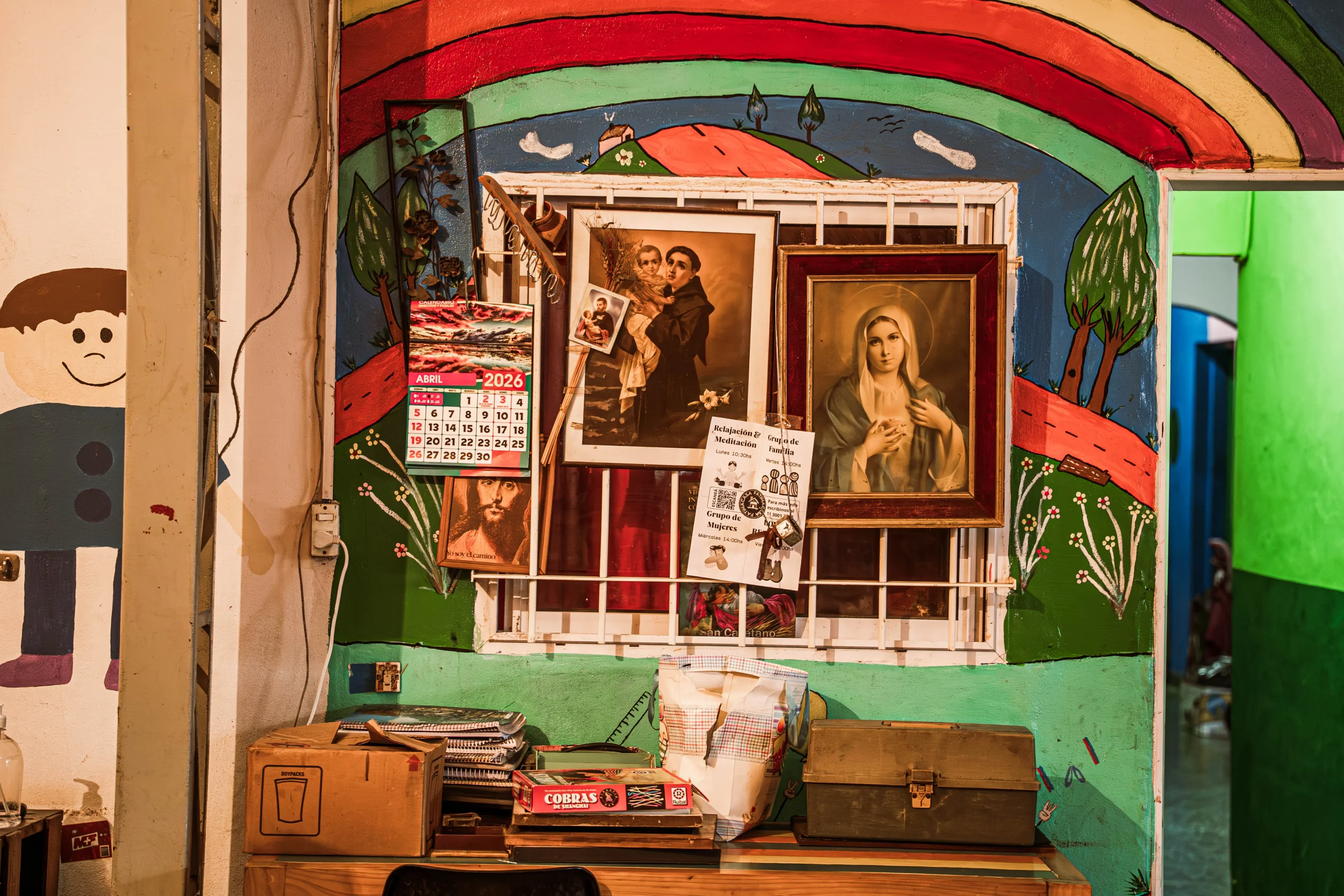Un rincón interior con una pared pintada con un mural colorido que representa un paisaje con un gran arco iris y árboles. Delante del mural hay una ventana enrejada donde se han colgado dos cuadros religiosos (uno de San Antonio con el Niño Jesús y otro de la Virgen María), un calendario de abril de 2026 y otros papeles. Abajo hay una mesa con cajas de cartón, útiles y un baúl metálico.