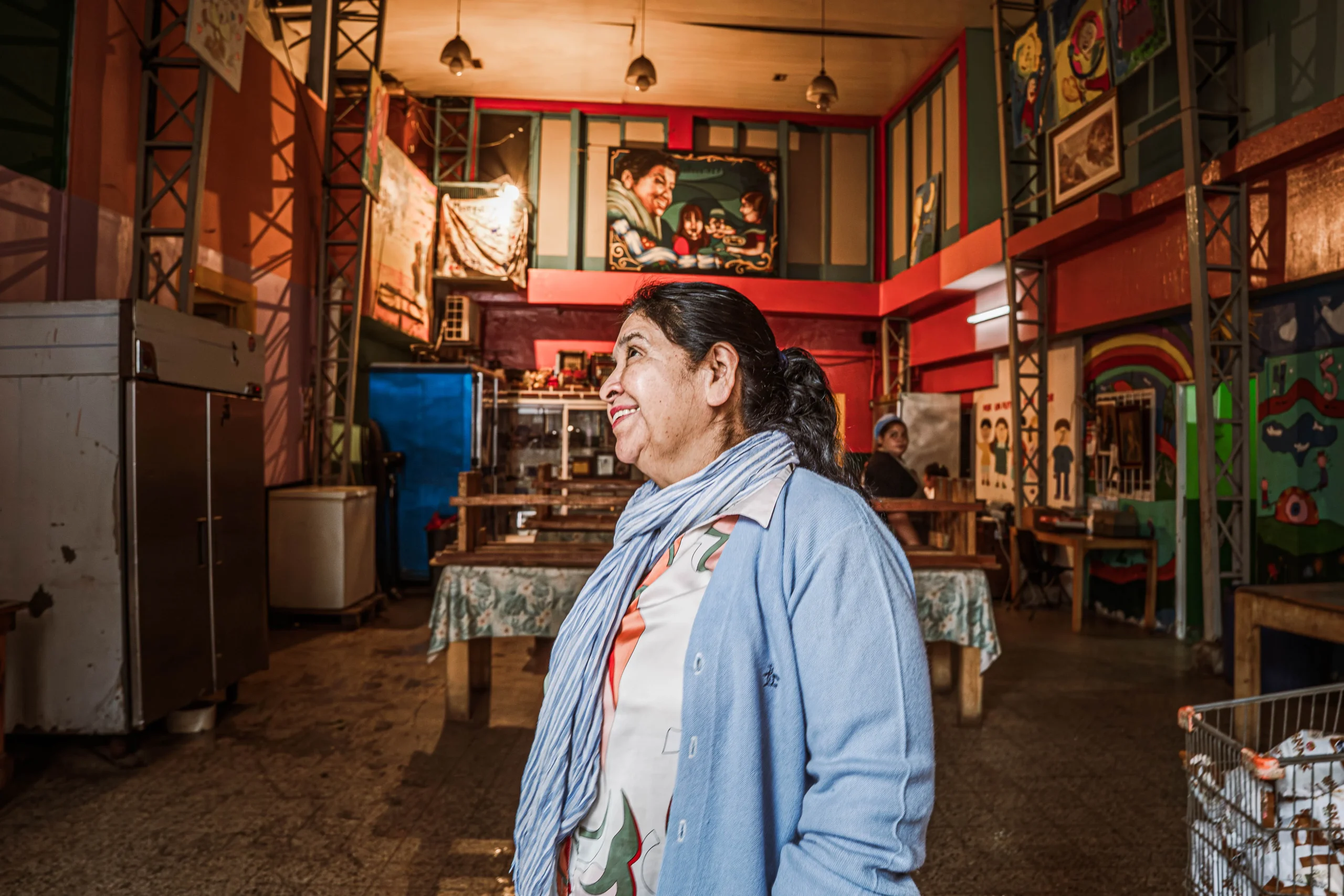 Una mujer de mediana edad con cabello oscuro recogido sonríe cálidamente mientras mira hacia un lado. Viste una chaqueta de punto azul claro y un pañuelo estampado. Está de pie en un gran espacio interior, posiblemente un comedor comunitario, que tiene techos altos, estructuras metálicas, mesas largas de madera con manteles, neveras industriales y un mural colorido en la pared del fondo.