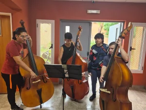 Tres jóvenes músicos de pie practican el contrabajo sosteniendo sus arcos y leyendo una partitura en un atril central, mientras un cuarto joven los observa a su lado en un salón de paredes color terracota.