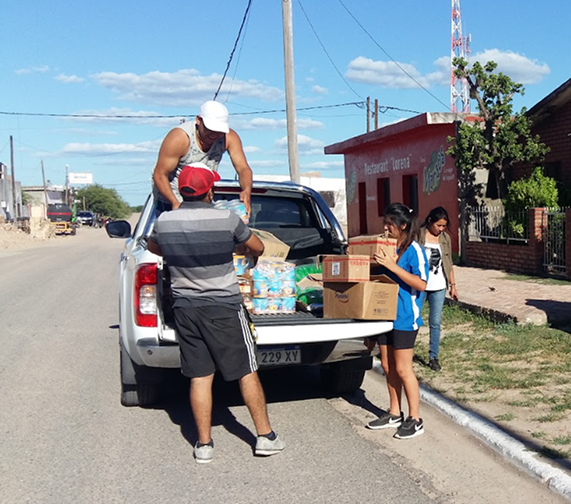 Un grupo de personas descarga cajas y paquetes de alimentos de la caja de una camioneta blanca estacionada en una calle de un barrio residencial. Un hombre desde arriba del vehículo entrega suministros a otro hombre y a una joven que esperan abajo para recibirlos. Al fondo se ve una casa de color rojo y un día despejado.