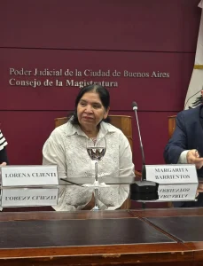 Margarita Barrientos sentada en una mesa de conferencias durante un evento en el Consejo de la Magistratura del Poder Judicial de la Ciudad de Buenos Aires. Viste una blusa blanca calada y está ubicada frente a un cartel con su nombre y un micrófono. De fondo, una pared color borgoña exhibe en letras plateadas el nombre de la institución. La imagen captura un momento de diálogo institucional.