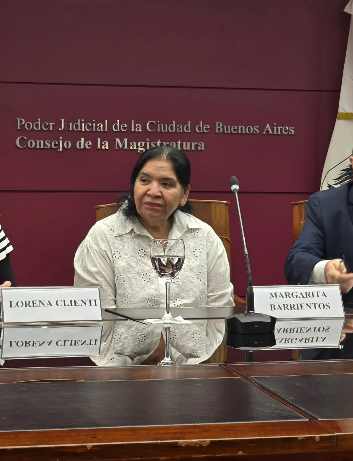 Margarita Barrientos sentada en una mesa de conferencias durante un evento en el Consejo de la Magistratura del Poder Judicial de la Ciudad de Buenos Aires. Viste una blusa blanca calada y está ubicada frente a un cartel con su nombre y un micrófono. De fondo, una pared color borgoña exhibe en letras plateadas el nombre de la institución. La imagen captura un momento de diálogo institucional.