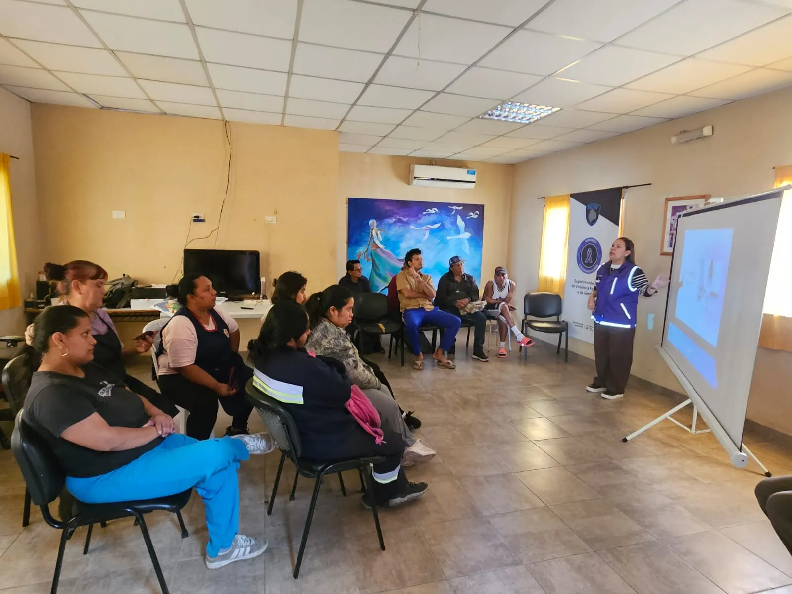 Una mujer con un chaleco institucional azul brinda una charla o capacitación a un grupo de personas sentadas en una sala. La mujer señala una pantalla de proyección mientras el público escucha atentamente. En el salón se observa un banner de la Superintendencia de Violencia Familiar y de Género de la Policía, un mural artístico al fondo y ventanas con cortinas amarillas.