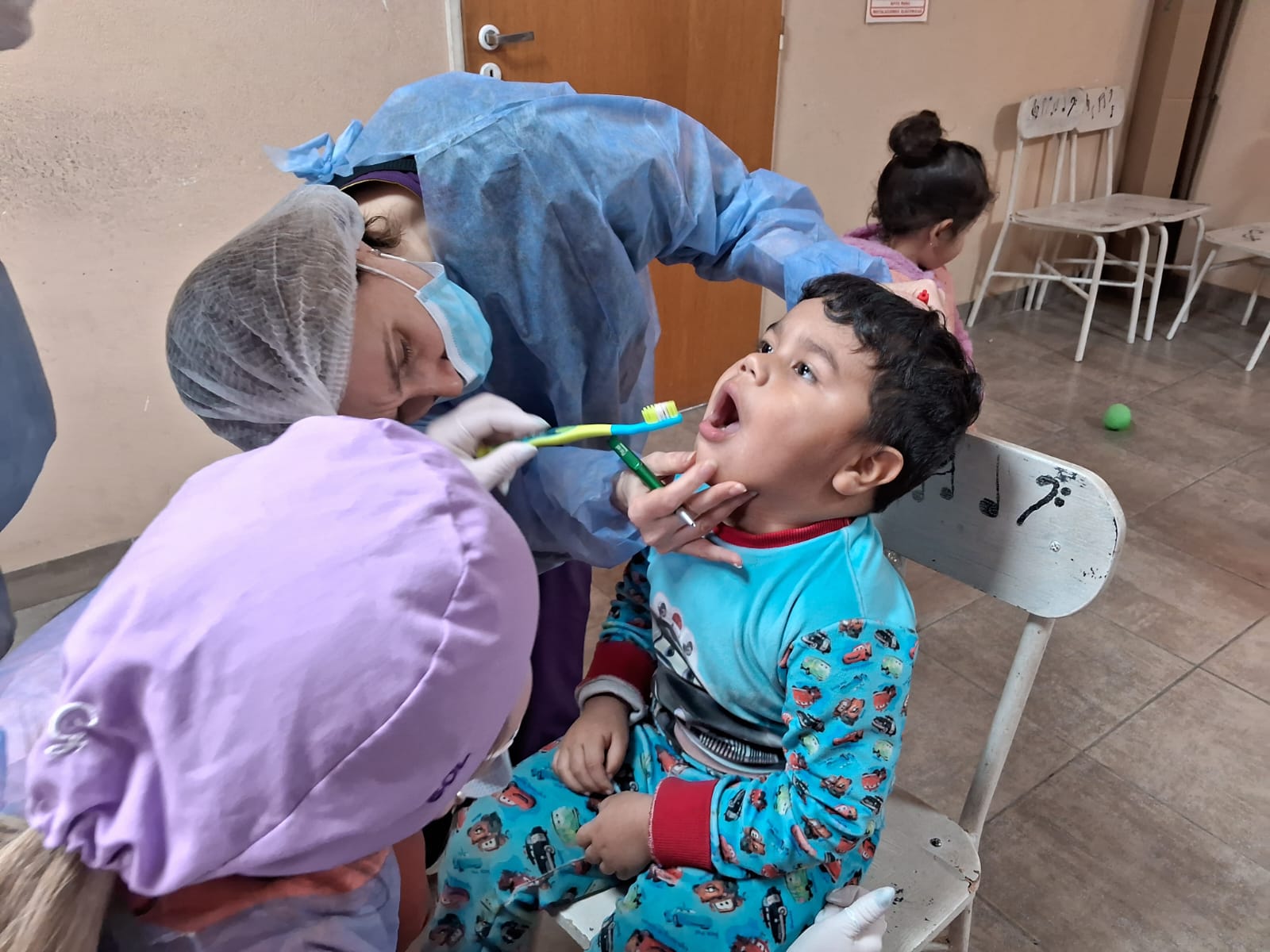 Niño menor de tres años con la boca abierta reclinado sobre una silla, atendido por dos médicas odontológicas que le controlan su higiene bucal