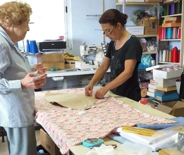 Una mujer con anteojos trabaja en un taller de costura colocando un molde de papel sobre una tela estampada extendida en una mesa grande, mientras una mujer mayor la observa de perfil a su lado en un entorno con máquinas de coser y estanterías llenas de hilos.