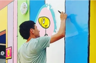 Un joven de espaldas, vestido con una remera gris, dibuja con un marcador negro sobre un mural colorido en una pared exterior. El mural tiene franjas verticales de colores vibrantes como verde, celeste, rosa y amarillo, e incluye la ilustración de un personaje esquemático con cabeza amarilla dentro de una forma circular.