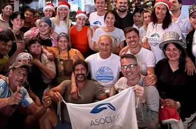 Un grupo numeroso de personas posa sonriente para una foto grupal en un ambiente festivo. Algunos integrantes llevan gorros de Papá Noel y remeras blancas con el logo de 