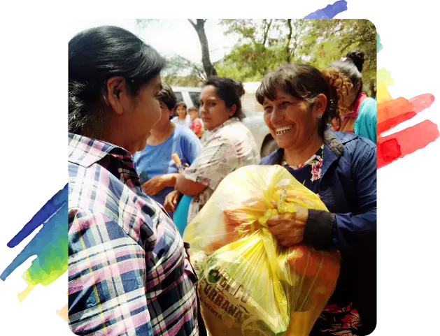 Dos mujeres, vestidas con camisas de cuadros, se sonríen y sostienen una bolsa de tela de color amarillo que contiene alimentos en una reunión comunitaria. Al fondo hay otros residentes esperando en una fila. Esta fotografía se superpone con un diseño artístico de pinceladas de pintura de los colores del arco iris con un fondo negro
