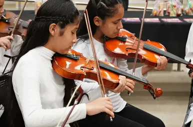 Dos niñas concentradas tocan el violín durante una presentación musical. Ambas visten remeras blancas de manga larga y sostienen sus instrumentos con técnica, mientras utilizan sus arcos sobre las cuerdas. Al fondo, se alcanza a ver una mesa con folletos y lo que parecen ser pequeños obsequios o refrigerios para el evento.