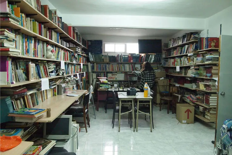 Una vista del interior de una habitación llena de estanterías repletas de libros en las paredes izquierda y derecha. Hay una persona de pie en el centro de la sala, de espaldas a la cámara. Un conjunto de mesas y sillas de estudio se encuentran en el centro. La habitación está iluminada por luz natural que entra a través de ventanas en la pared del fondo. Una puerta se encuentra en la pared de la derecha.