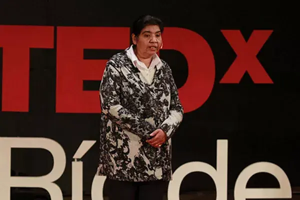 Margarita Barrientos parada en una escenario vestida con una camisa con flores. De fondo se ve el cartel de charlas TED. La imagen captura un momento de exposición de Margarita Barrientos en las Charlas TED contando sobre su experiencia y trayectoria de la fundación.