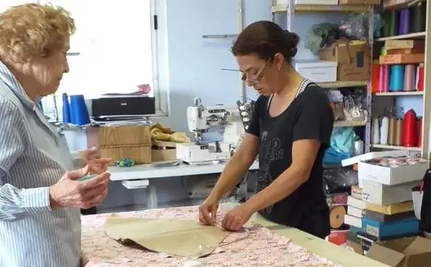 Una mujer con anteojos trabaja en un taller de costura colocando un molde de papel sobre una tela estampada extendida en una mesa grande, mientras una mujer mayor la observa de perfil a su lado en un entorno con máquinas de coser y estanterías llenas de hilos.