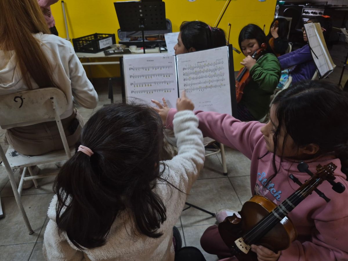 Dos niñas de espalda con cabello largo, tocan con sus dedos índices una partitura musical que está en un atril. Una de las niñas tiene en sus brazos un violín.