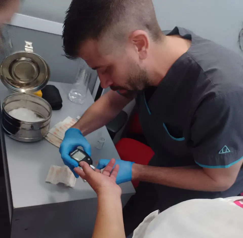 Un profesional de la salud con ambos guantes azules realiza una prueba de glucosa en sangre sosteniendo un glucómetro digital contra el dedo de una persona en un consultorio o sala médica.