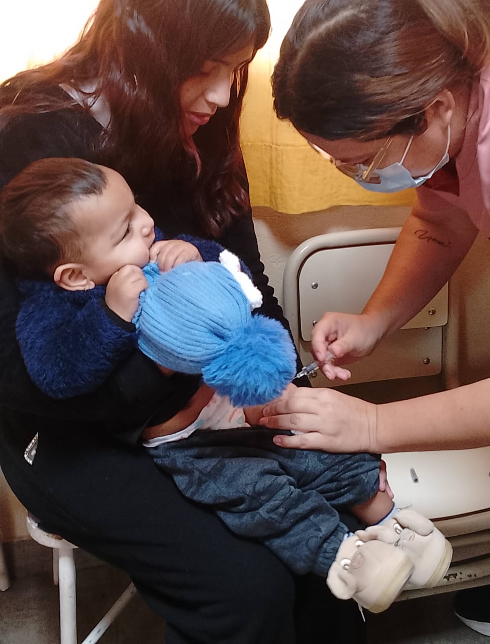 Un profesional de salud con barbijo aplica una vacuna a un bebe que es sostenido en brazos por una mujer sentada. El bebe se encuentra mordiendo un gorro celeste de lana.