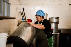 Mujer con cofia azul y delantal trabajando en una cocina industrial. Sostiene una olla metálica muy grande e inclinada. Al fondo, otras ollas de gran tamaño, utensilios y paredes de azulejos blancos.