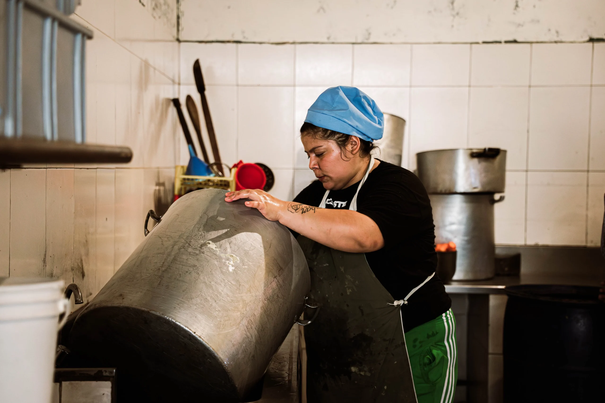 Mujer con cofia azul y delantal trabajando en una cocina industrial. Sostiene una olla metálica muy grande e inclinada. Al fondo, otras ollas de gran tamaño, utensilios y paredes de azulejos blancos.