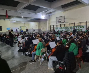 Una orquesta de jóvenes músicos ensaya en un gran salón con piso de baldosas y un techo de vigas de hormigón visto.