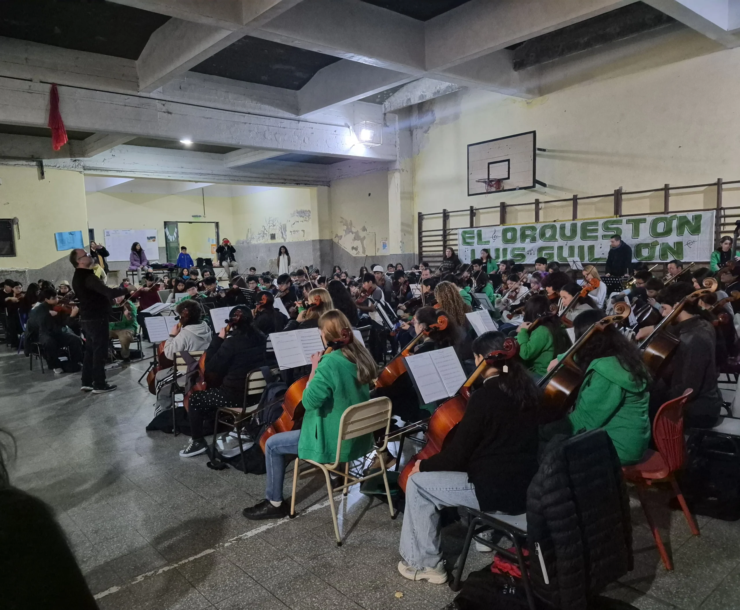Una orquesta de jóvenes músicos ensaya en un gran salón con piso de baldosas y un techo de vigas de hormigón visto.