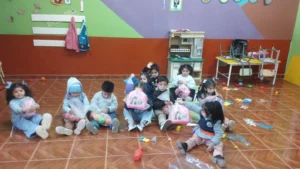 Grupo de niñas y niños pequeños sentados en el piso de un aula de jardín de infantes, sosteniendo juguetes y bolsas con regalos. El aula tiene paredes de colores, una cocinita de juguete y juguetes esparcidos por el suelo.