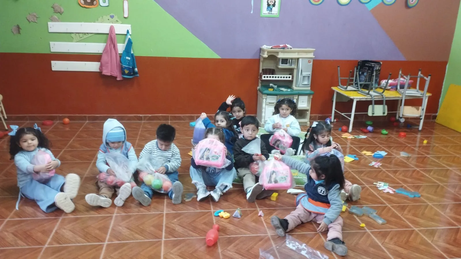 Grupo de niñas y niños pequeños sentados en el piso de un aula de jardín de infantes, sosteniendo juguetes y bolsas con regalos. El aula tiene paredes de colores, una cocinita de juguete y juguetes esparcidos por el suelo.