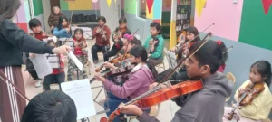 Integrantes de la Orquesta infanto juvenil Despertando Sueños ensayando: un grupo de niños y niñas tocan violines con sus atriles y partituras, dirigidos por una docente. El aula tiene paredes de colores y decoración infantil.