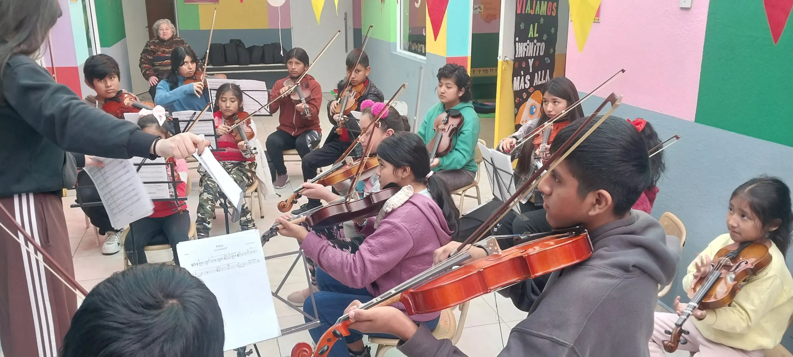 Integrantes de la Orquesta infanto juvenil Despertando Sueños ensayando: un grupo de niños y niñas tocan violines con sus atriles y partituras, dirigidos por una docente. El aula tiene paredes de colores y decoración infantil.