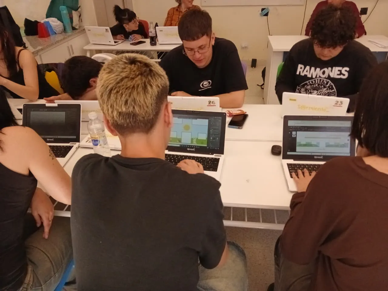 Grupo de jóvenes trabajando con notebooks alrededor de mesas blancas en un aula. En las pantallas se ve software de diseño. Están concentrados y hay botellas de agua y celulares sobre las mesas.