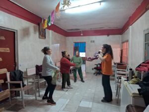 Foto de unas mujeres paradas en movimiento con pañuelos en la mano bailando. En el techo hay unas guirnaldas colgando y a los costados sillas.