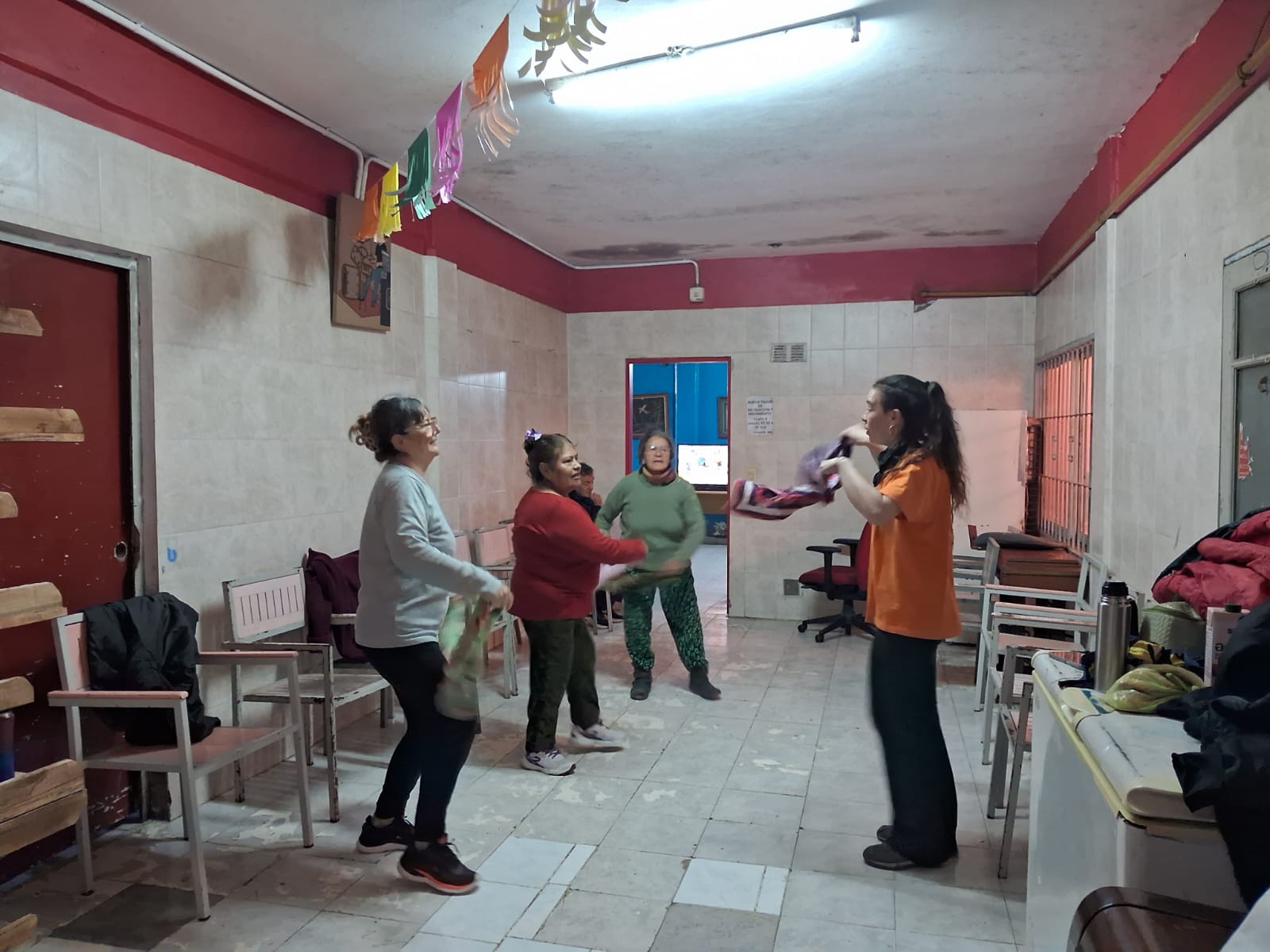 Foto de unas mujeres paradas en movimiento con pañuelos en la mano bailando. En el techo hay unas guirnaldas colgando y a los costados sillas.