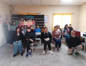 Foto grupal de unas quince personas jóvenes, en su mayoría mujeres, en un salón comunitario de piso de cerámica. Algunas están sentadas en sillas y otras de pie detrás. Al fondo hay un telón de tela negra pintado a mano con el cartel "La Pulpería" y siluetas de botellas y copas, junto a un panel de colores.