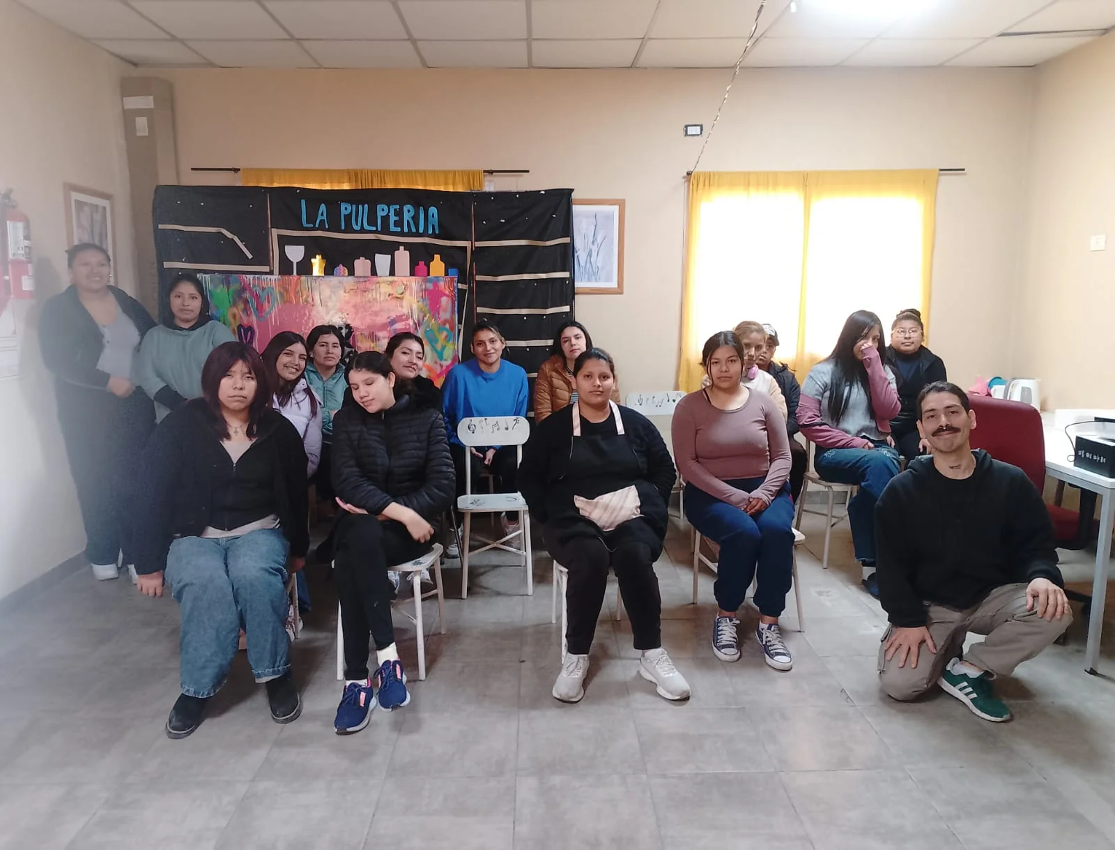 Foto grupal de unas quince personas jóvenes, en su mayoría mujeres, en un salón comunitario de piso de cerámica. Algunas están sentadas en sillas y otras de pie detrás. Al fondo hay un telón de tela negra pintado a mano con el cartel "La Pulpería" y siluetas de botellas y copas, junto a un panel de colores.