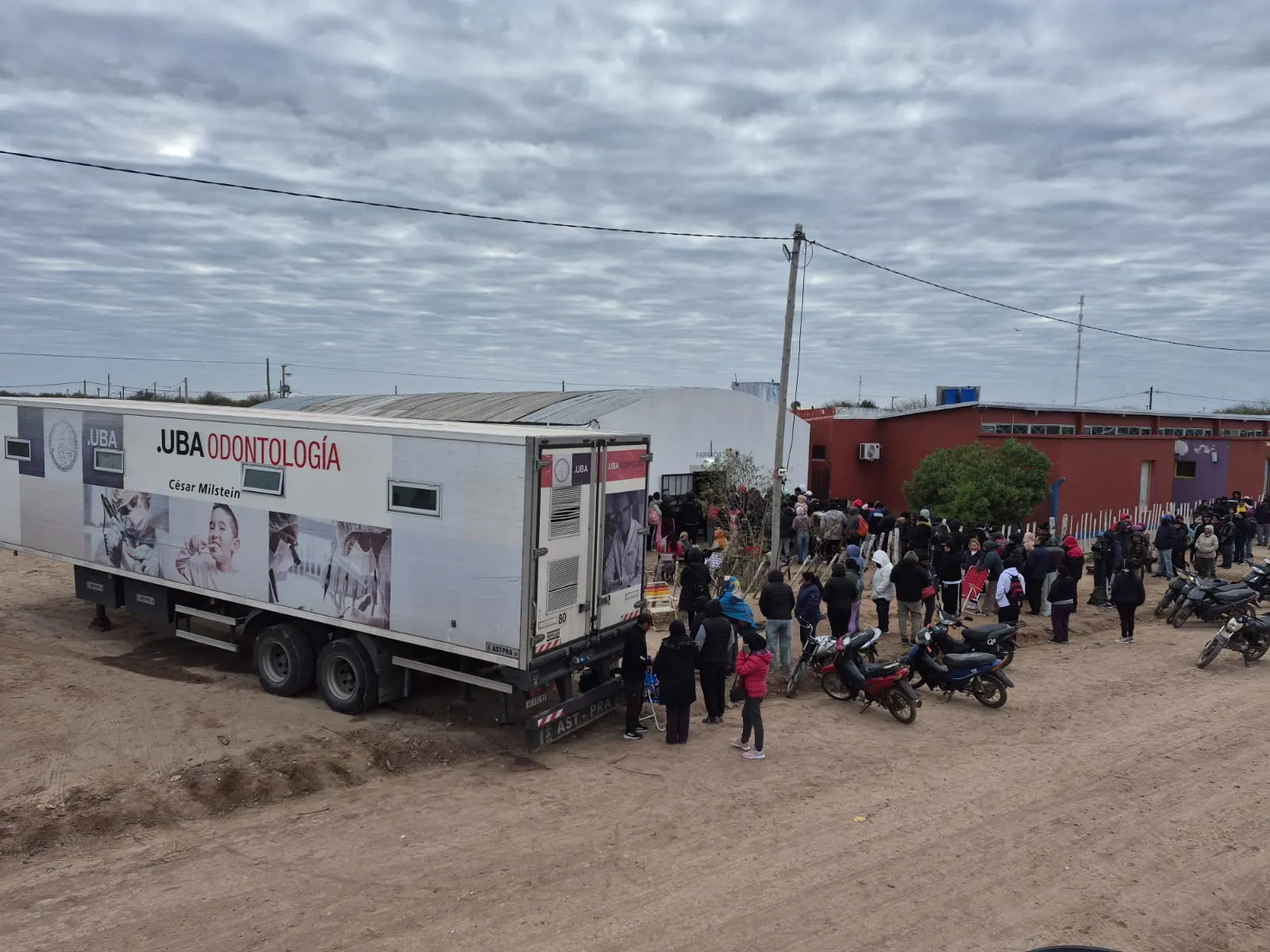 Camión-consultorio odontológico móvil de la UBA estacionado en una calle de tierra. Una multitud de personas hace fila frente a un edificio bajo. Alrededor hay motos estacionadas y el cielo está nublado.