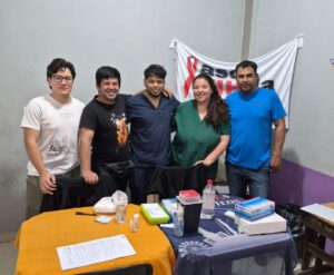 Grupo de cinco personas abrazadas mirando a la cámara de manera sonriente. Detrás una bandera blanca colgada y delante dos mesas con manteles