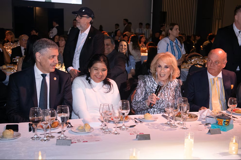 Mesa principal de una cena de gala. Cuatro invitados sentados tras un mantel blanco con copas, panes y velas. Una mujer de saco plateado con lentejuelas sostiene un micrófono mientras habla. Al fondo, el salón lleno de comensales.