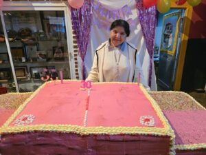 Persona sonriendo frente a una torta de cumpleaños muy grande de color rosa sobre una mesa