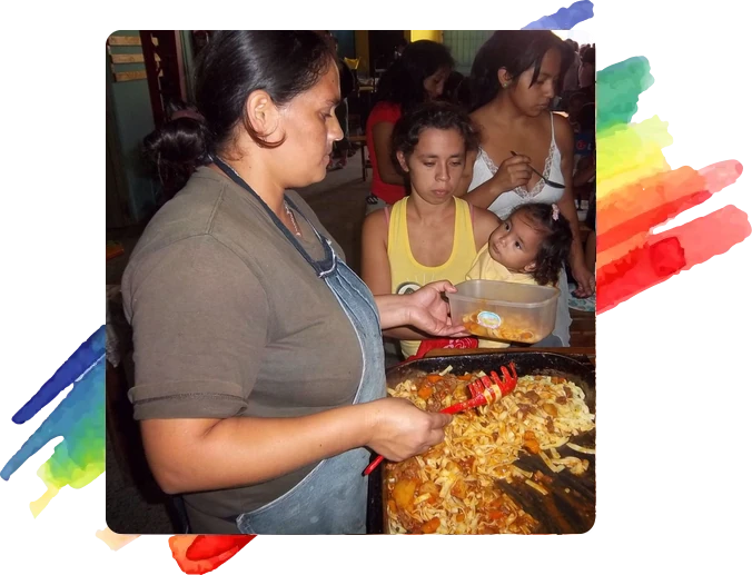 Mujer con delantal sirviendo comida recién preparada desde una gran sartén a un recipiente plástico. Detrás, varias personas esperan su porción, entre ellas una mujer que sostiene a una niña pequeña en brazos