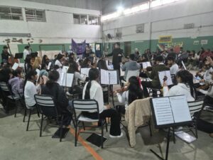 Una orquesta de jóvenes músicos ensaya en un gran salón con piso de baldosas y un techo de vigas de hormigón visto.